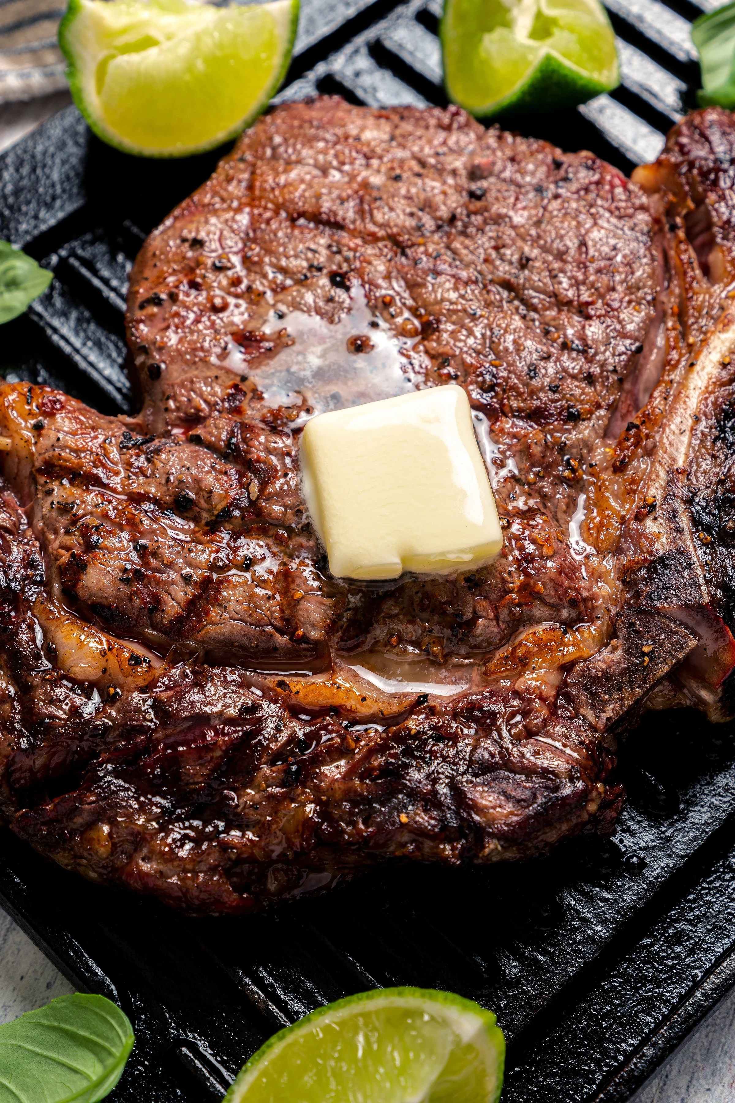 Macro food styling of a grilled ribeye steak with a melting square of gourmet butter. Dramatic studio lighting captures the rich sear marks, glossy juices, coarse black pepper, and bright green lime garnishes for a food ad.
