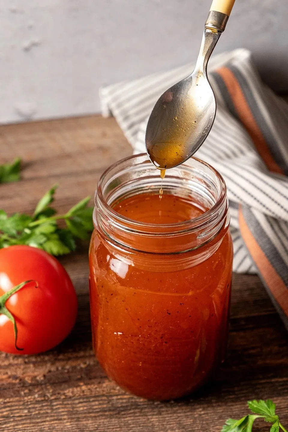 Commercial product photography of Gertie's Salad Dressing. A silver spoon drips a zesty orange-red vinaigrette over an open mason jar on a rustic wood surface styled with a fresh tomato and parsley.
