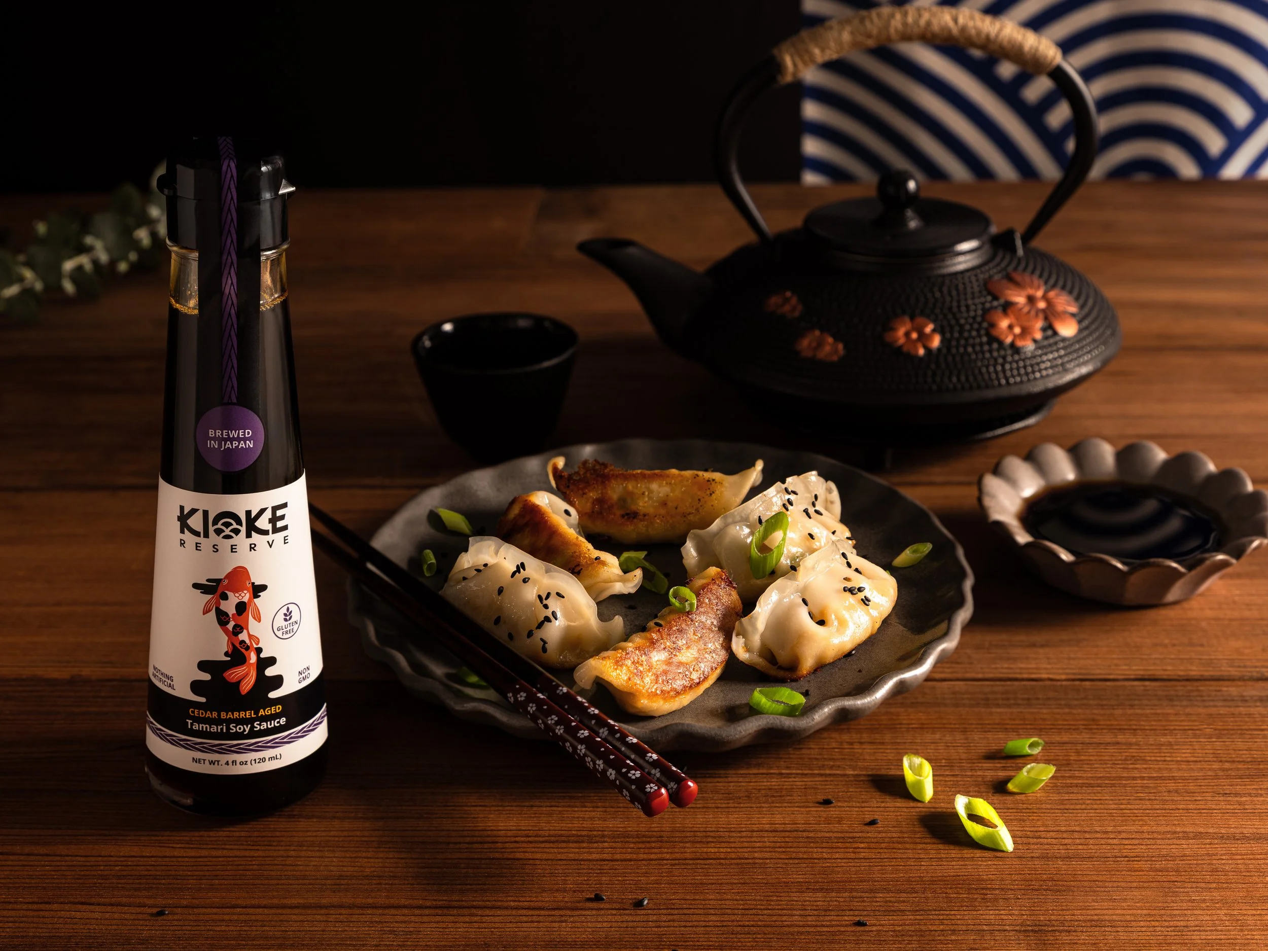 Editorial shot of a premium Japanese tamari soy sauce bottle paired with a fresh dumpling dinner presentation. Artistically composed on a warm wood surface with green onions and a small dipping dish for a condiment brand.