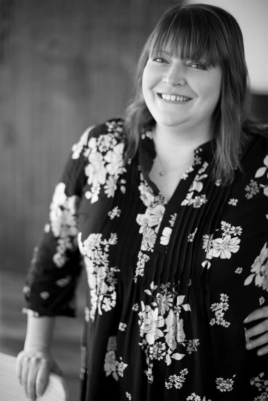 Black and white professional headshot portrait of a smiling woman with bangs wearing a floral print blouse, styled for a creative studio about me page.