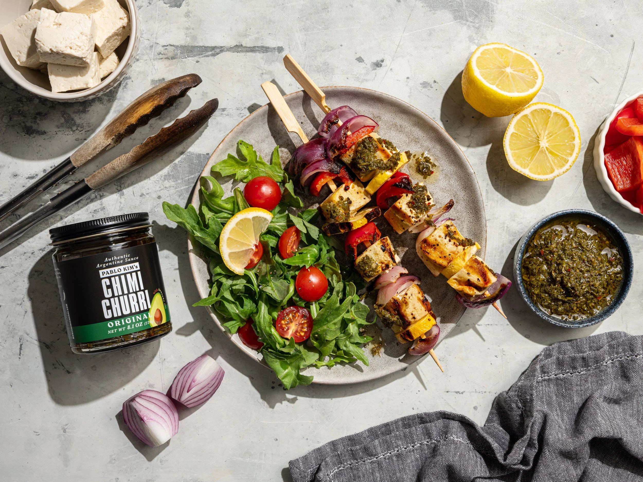 Overhead food photography of grilled vegetarian tofu kebabs. High-contrast direct sunlight casts sharp shadows, emphasizing charred skewers, fresh green herbs, cherry tomatoes, and lemons for a premium CPG food campaign.