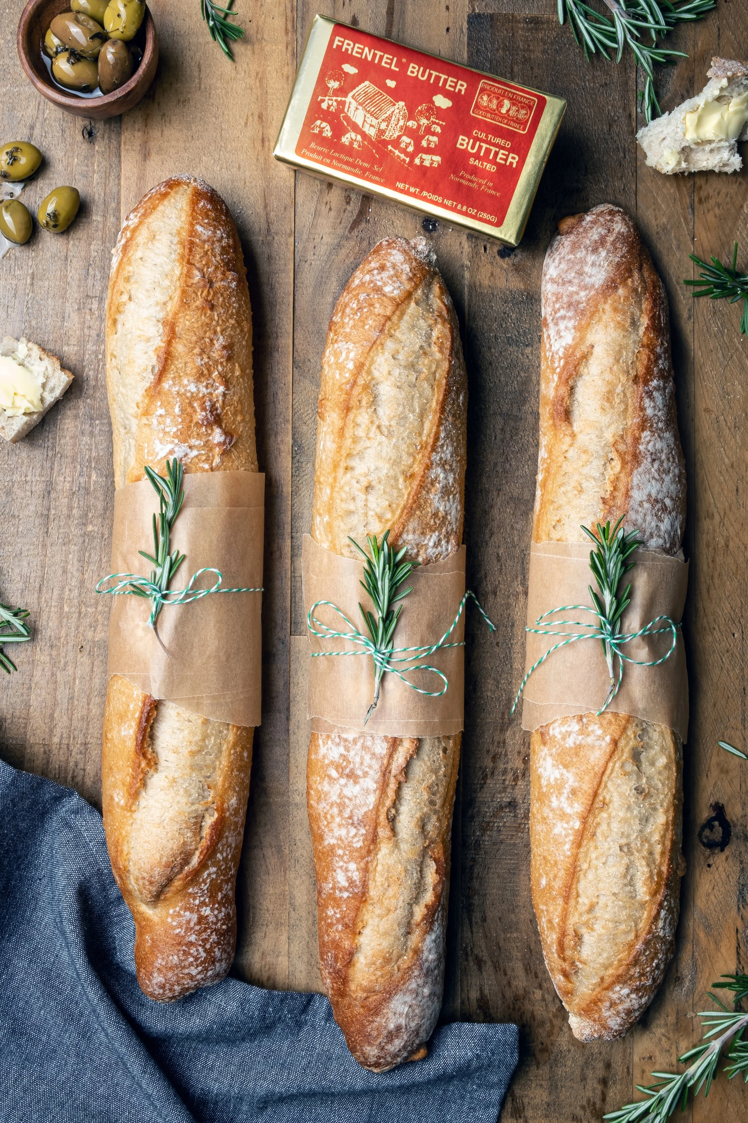 Premium editorial flat lay capturing a rustic bakery aesthetic. Top-down studio lighting highlights flour-dusted baguettes, a blue linen napkin, fresh rosemary needles, and luxury butter packaging for a CPG campaign.