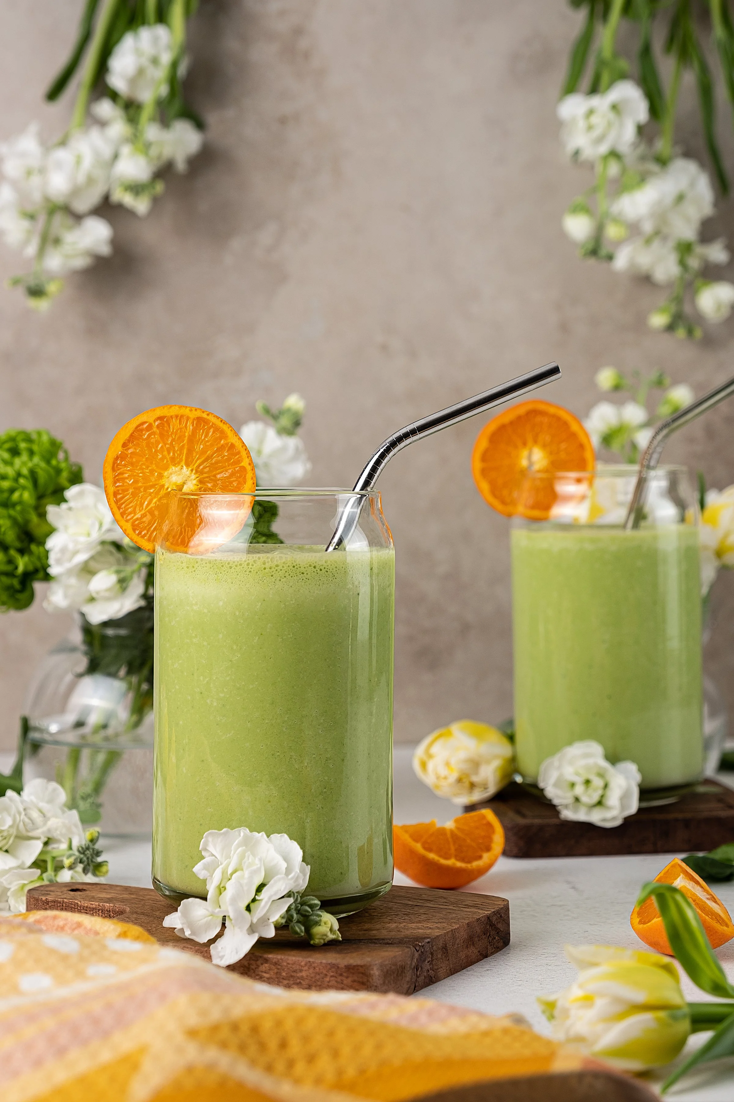 Commercial beverage photography for Dah! Lassi. Two green probiotic yogurt smoothie drinks in can-shaped glasses are styled on a wood coaster with fresh orange wheels, metal straws, and white flowers on a neutral backdrop