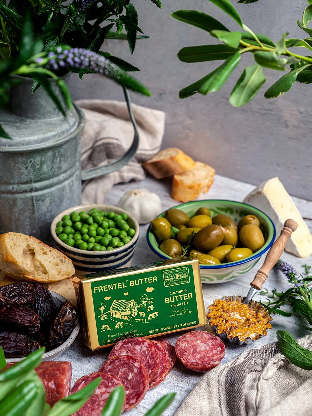 Moody commercial food photography of a charcuterie spread featuring a green package of Frentel Unsalted Butter, sliced salami, whole grain mustard, green olives, fresh peas, and artisanal baguette slices.