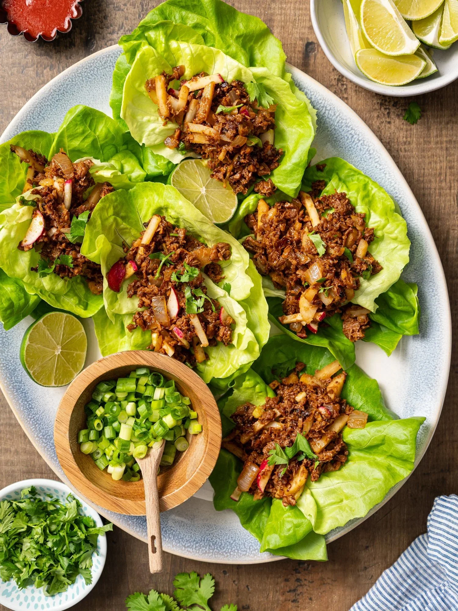 Overhead food photography of healthy vegetarian lettuce wraps on a large platter. Crisp butter lettuce cups are filled with a savory plant-based mixture, styled with fresh lime wedges and chopped green onions.