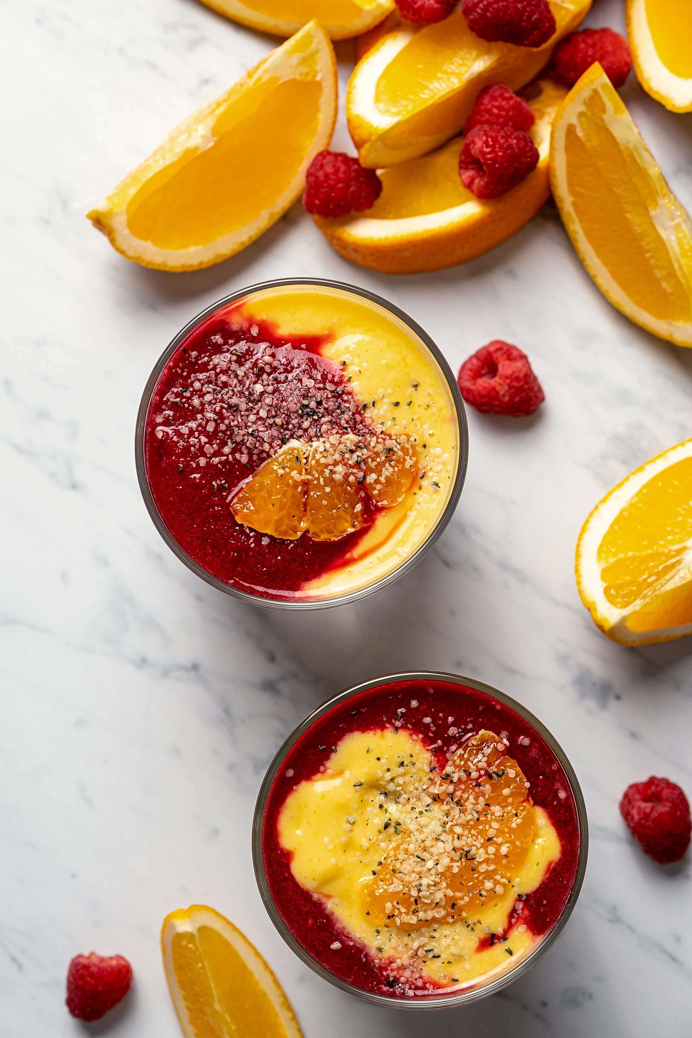 Commercial beverage photography for Dah! featuring a flat lay of two layered fruit smoothie bowls. Styled with swirls of orange citrus and pink raspberry purées, fresh orange slices, berries, and chia seeds on marble.