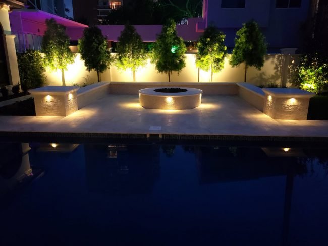 Outdoor patio with seating and fire pit, illuminated by warm lights, overlooking a pool at night.