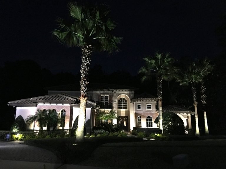 A large, well-lit house at night with palm trees in the foreground.