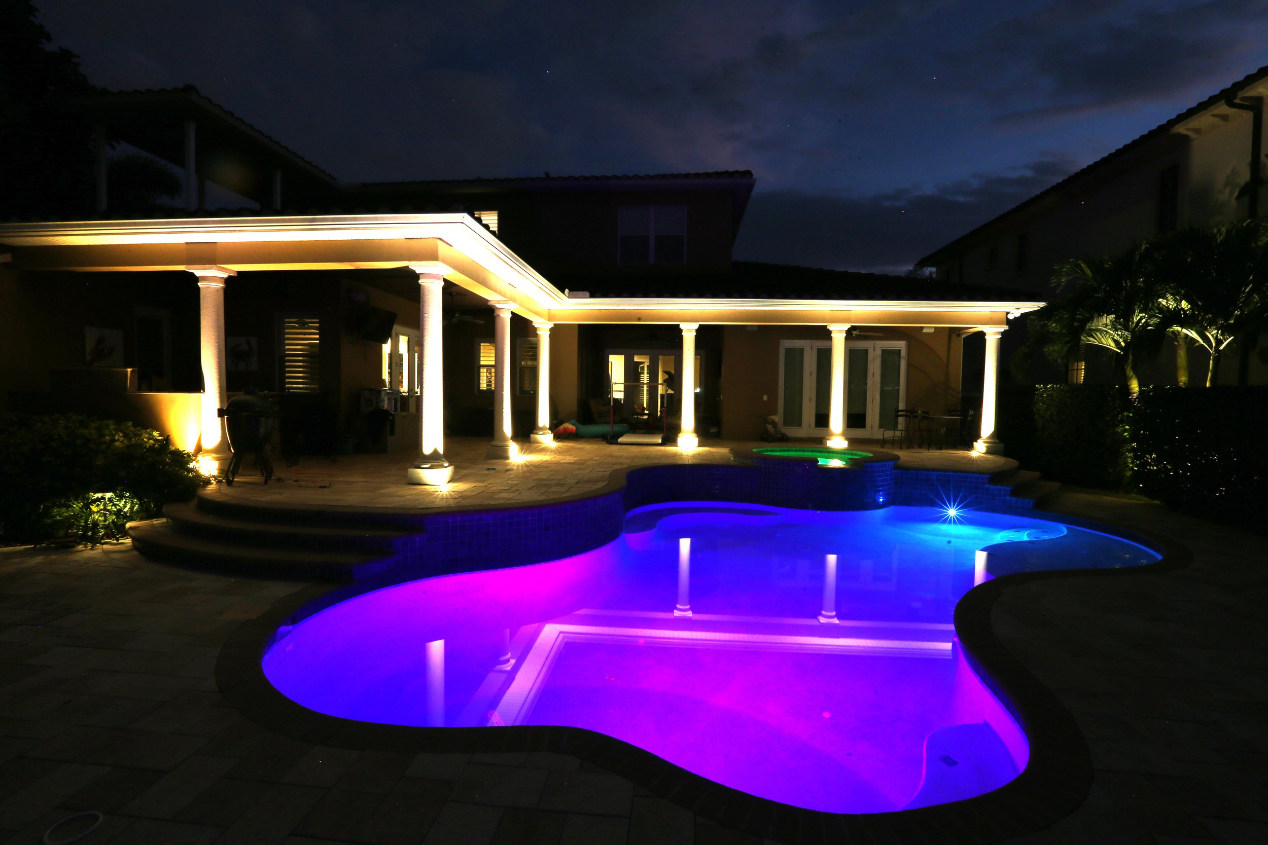 Backyard swimming pool with colorful LED lighting, surrounded by a patio and columns, at night. Lighting install by signature lighting & electrical, Tampa Florida