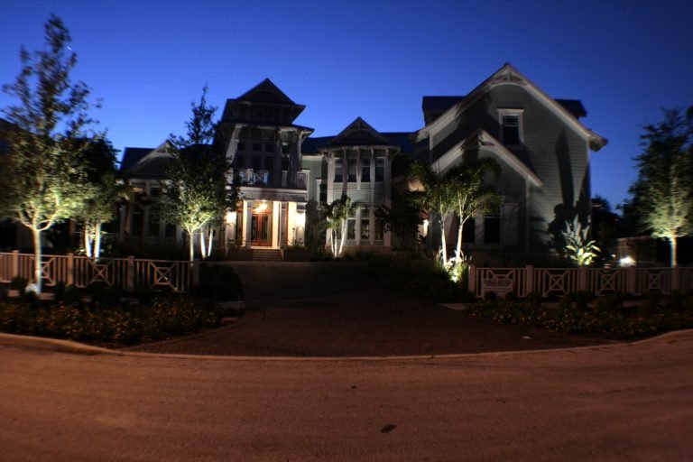 A large, well-lit two-story house with multiple gables and a front porch, surrounded by trees and illuminated at night.