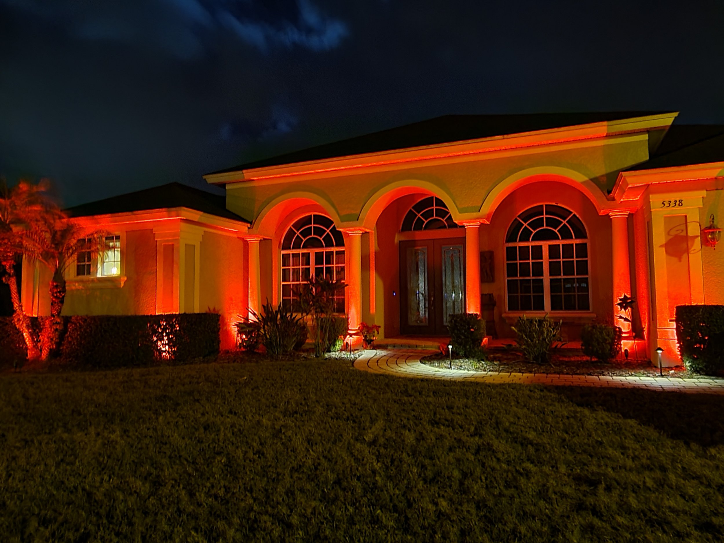 House with front yard illuminated by red lights at night, featuring arched windows and columns.