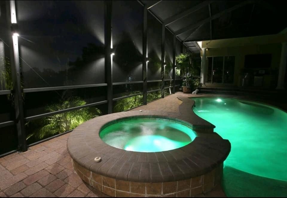 Enclosed swimming pool and hot tub area, night, illuminated with green lights, patio, and tropical plants.