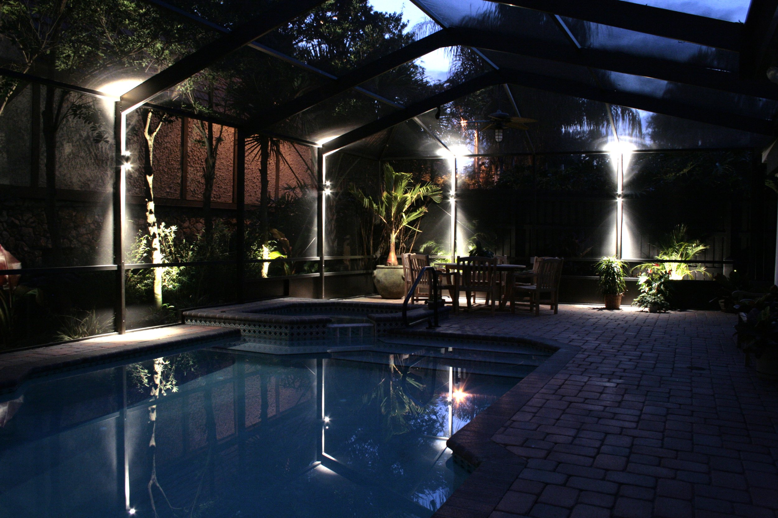 Screened-in patio with pool, plants, and outdoor furniture at night.