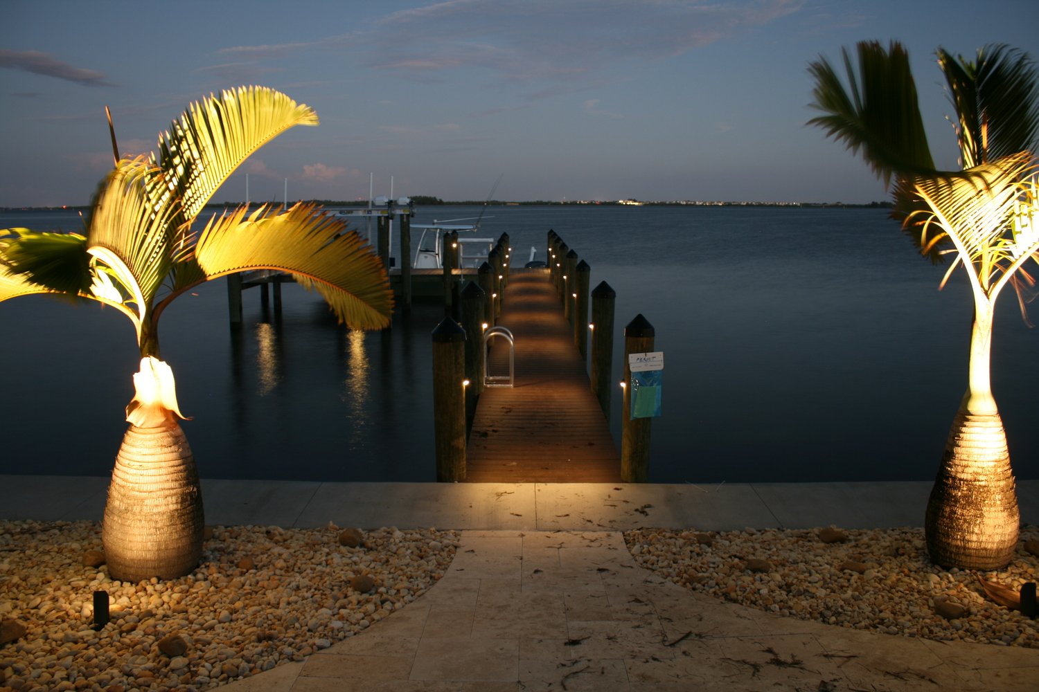 A wooden dock extends into a calm body of water at dusk, framed by illuminated palm trees on either side. The sky is a deep blue with subtle cloud formations, creating a tranquil and inviting atmosphere.