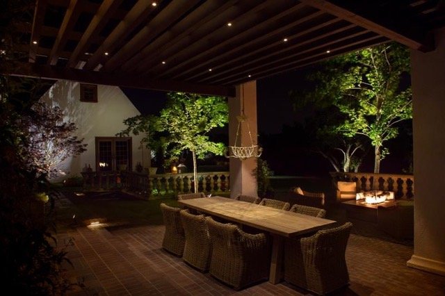 A dimly lit outdoor patio features a long wooden dining table with wicker chairs. The area is covered by a pergola with small lights. Trees and a house are visible in the background, creating a serene evening ambiance.