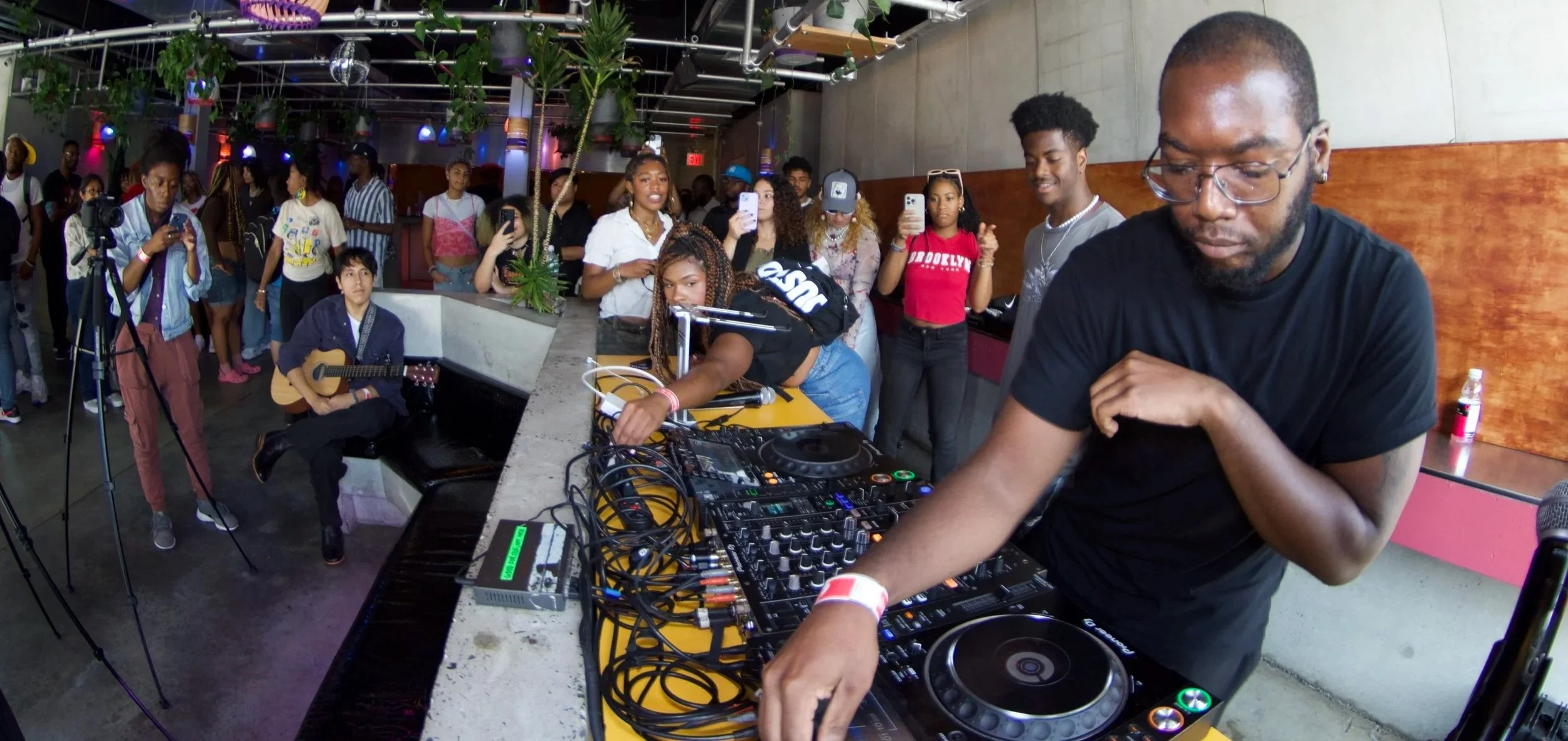 DJ performing with turntables and mixer at an indoor event, with a crowd of young people watching and recording, one person playing guitar, and others capturing the moment on their phones.