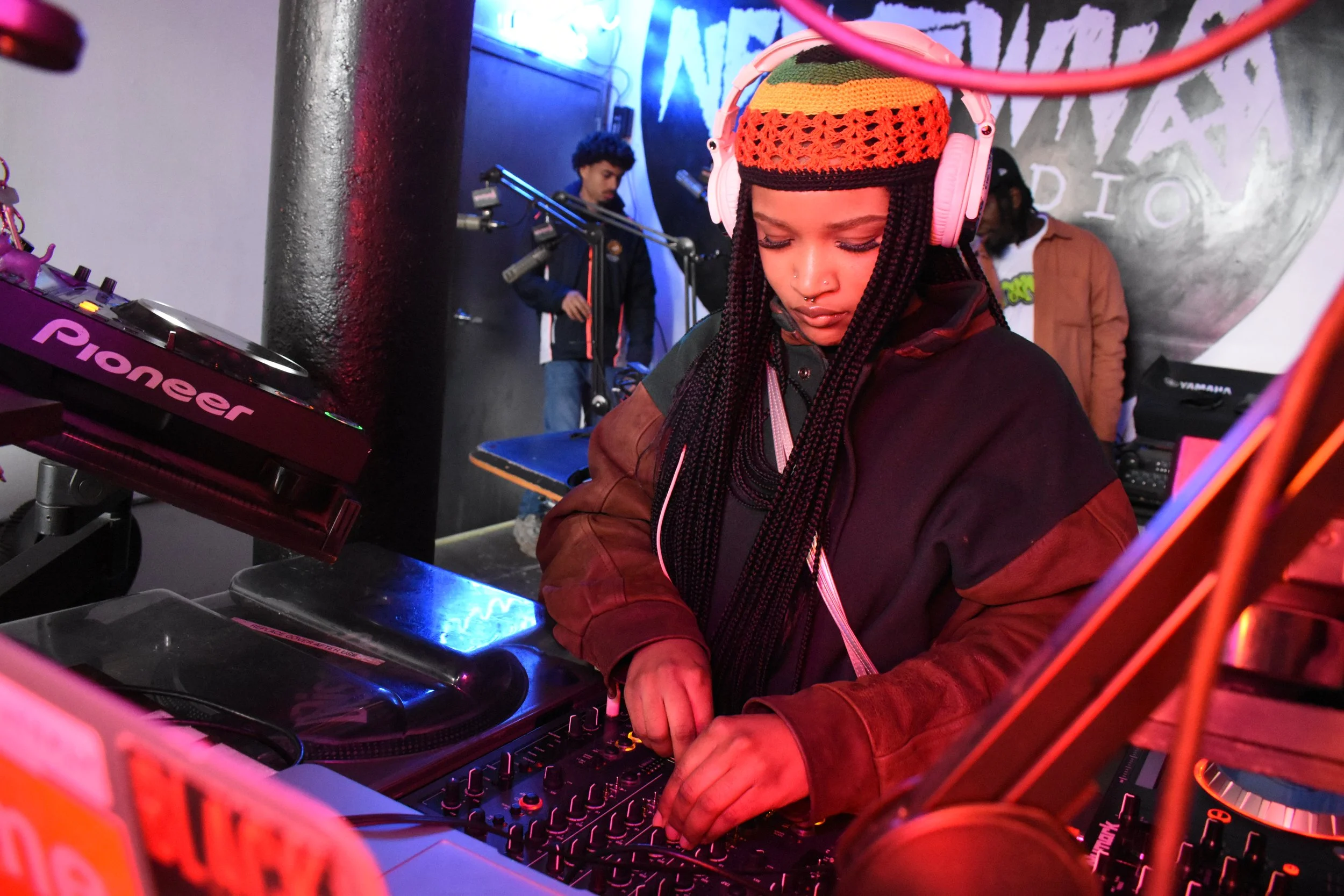 A female DJ with long braided hair, wearing headphones and a colorful crocheted hat, is adjusting controls on a DJ mixer in a club or music venue, with other people and DJ equipment in the background.