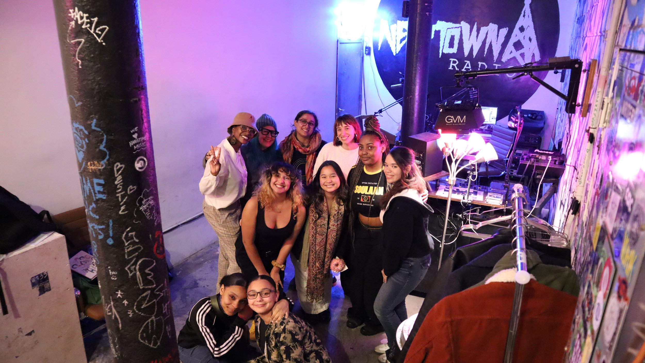 Group of ten women smiling and posing for a photo in a colorful, graffiti-decorated club or event space with DJ equipment in the background.