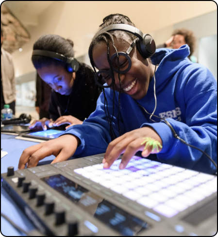 Two young women using electronic music equipment, one smiling and wearing headphones, in a casual indoor setting.