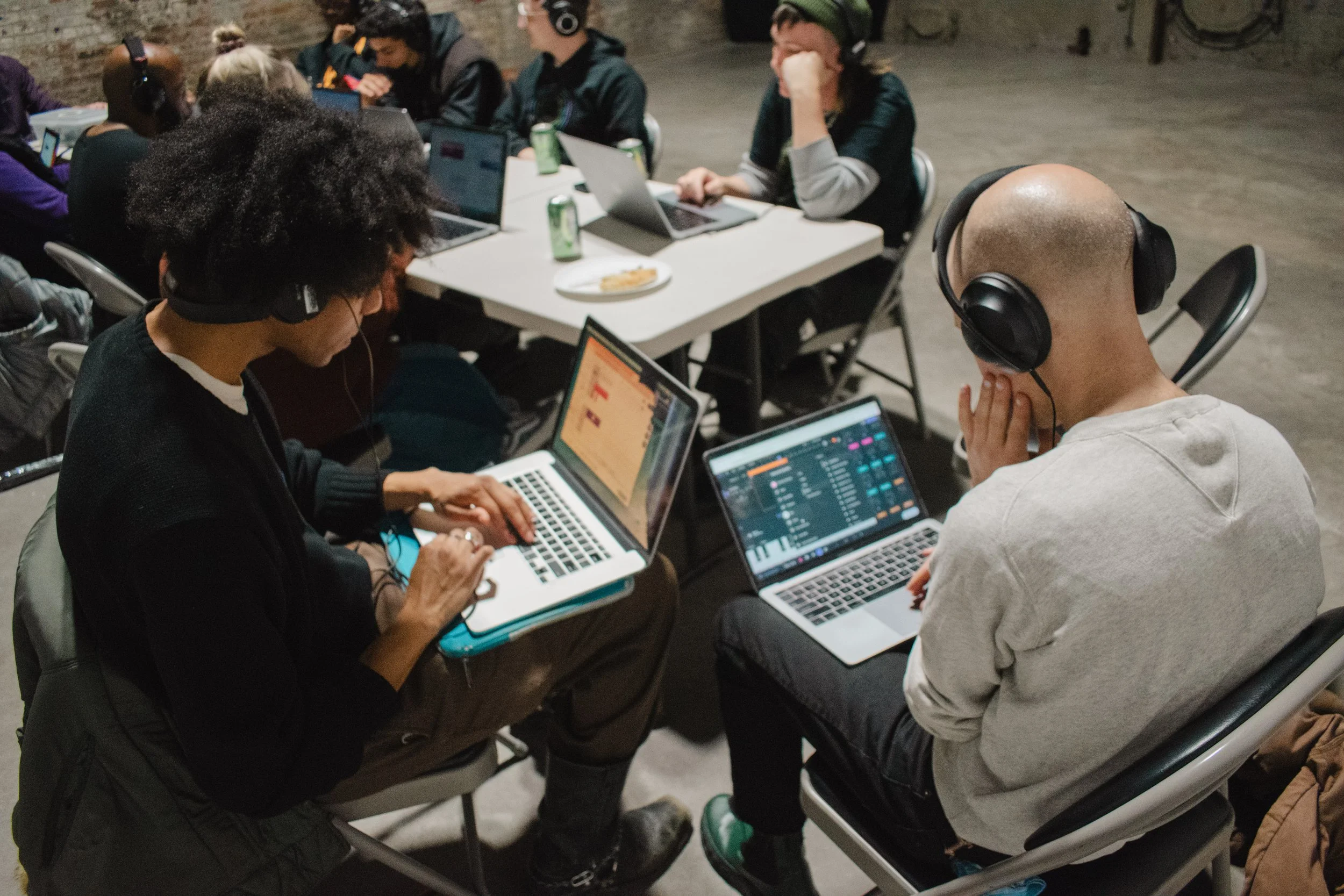 Group of people sitting around a long table working on laptops in a dimly lit room, some wearing headphones, with drinks and snacks on the table.
