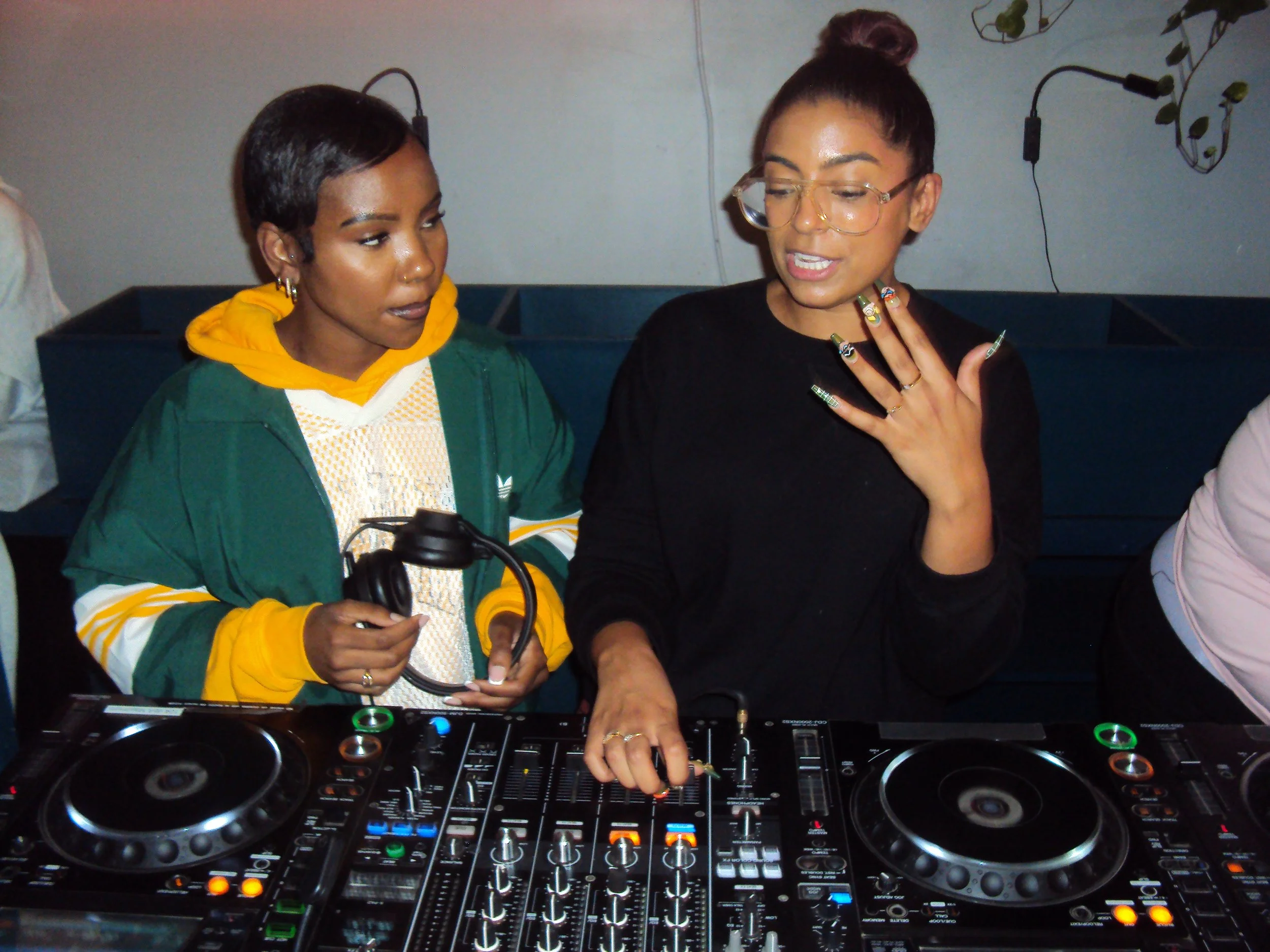 Two women at a DJ booth, one wearing a green and yellow jacket with headphones, the other with glasses and black shirt, gesturing while speaking.