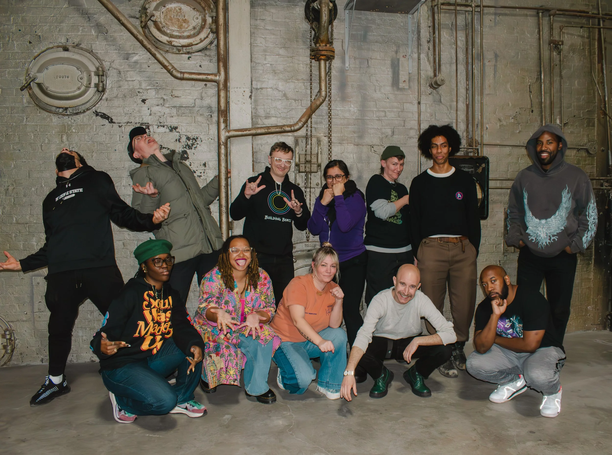 Group of diverse people posing and making various expressions in an industrial space with exposed bricks and pipes.