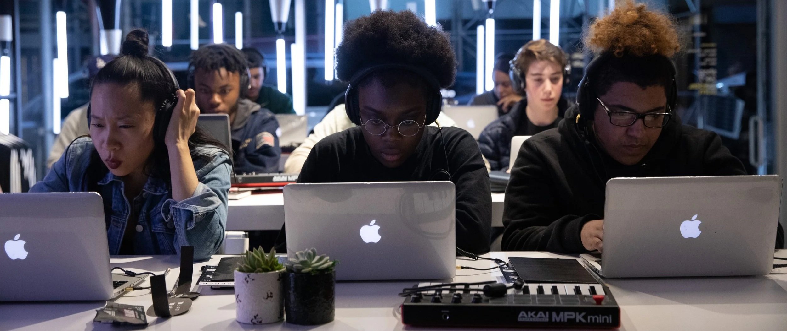 Group of young people wearing headphones working on Apple MacBook laptops at a tech event or workshop.