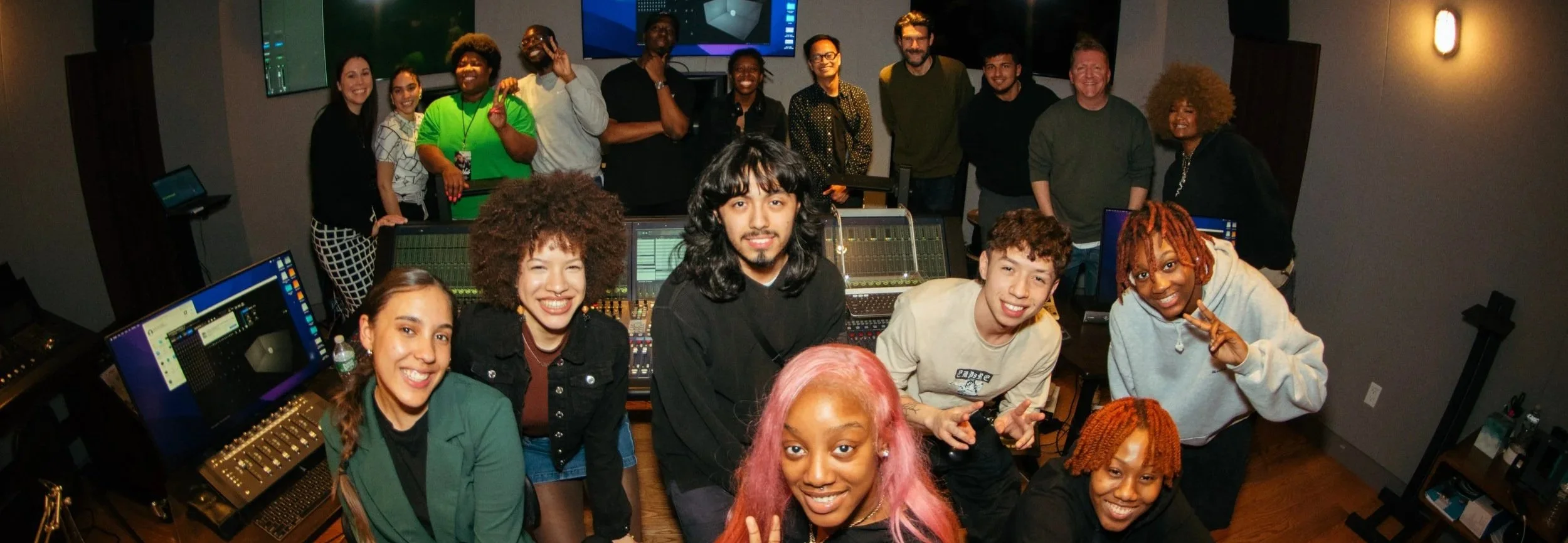 A diverse group of people in a recording studio, smiling and posing for a group photo. Several people are in front of sound mixing equipment and computers, with others standing behind them against a wall with monitors.