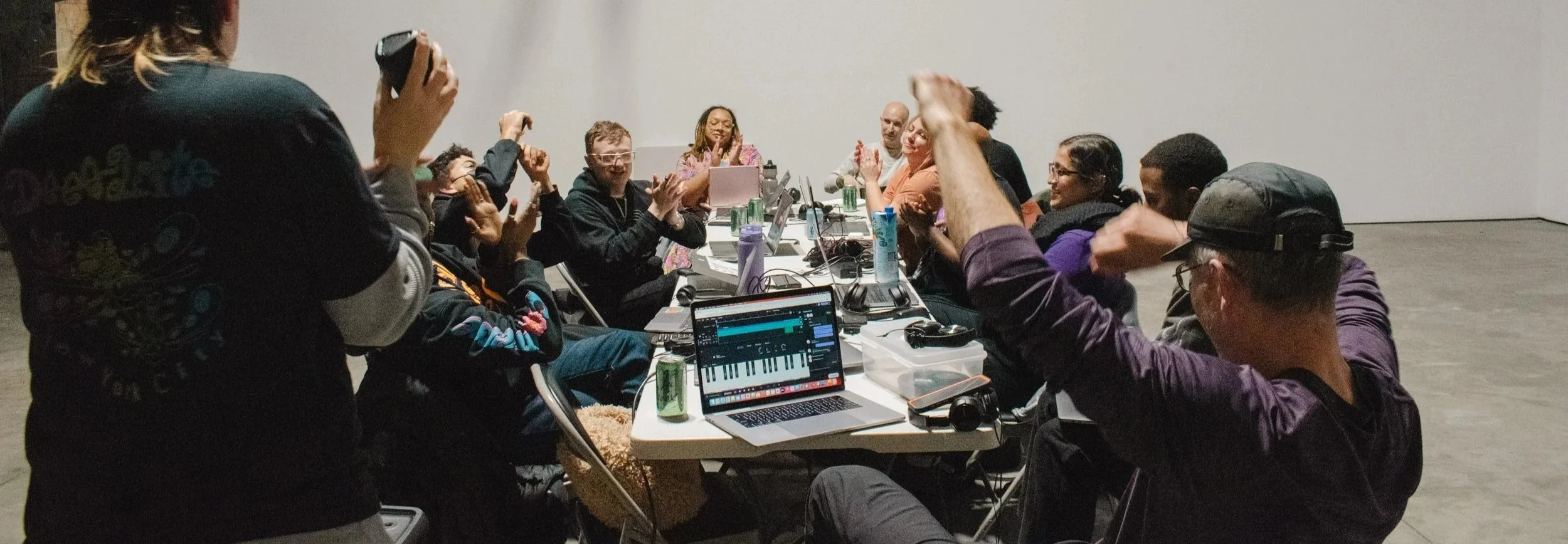 A diverse group of people gathered around a long table, some clapping and others raising their hands, with laptops and drinks on the table, in a room with plain white walls.