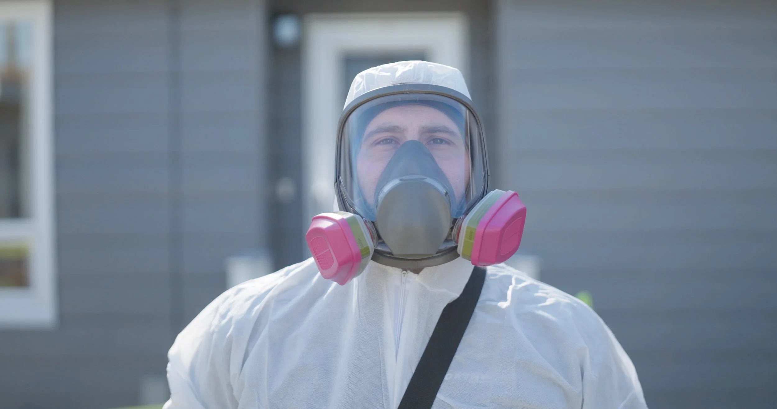 Person outdoors wearing full protective PPE, including a white suit, a face shield, and a respirator mask with pink filters