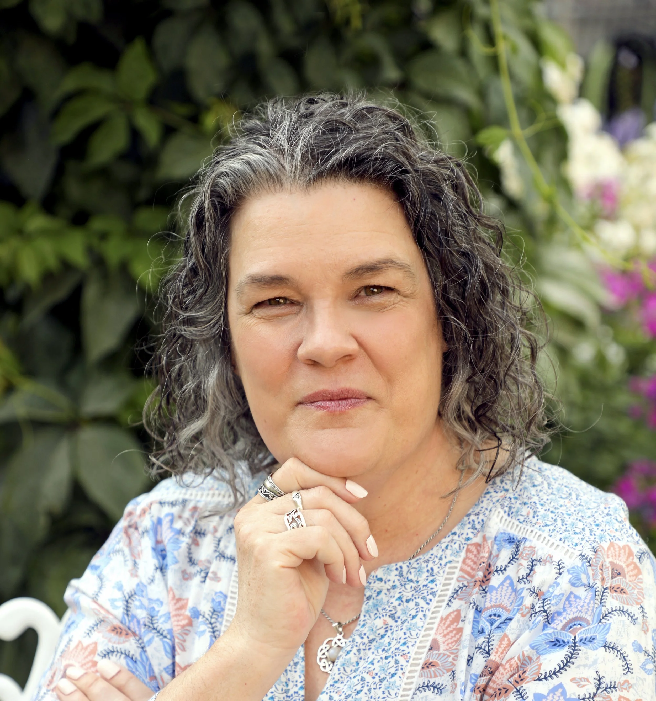 Portrait of a woman with wavy hair in a garden, wearing a patterned blouse and silver jewelry, with a thoughtful expression.