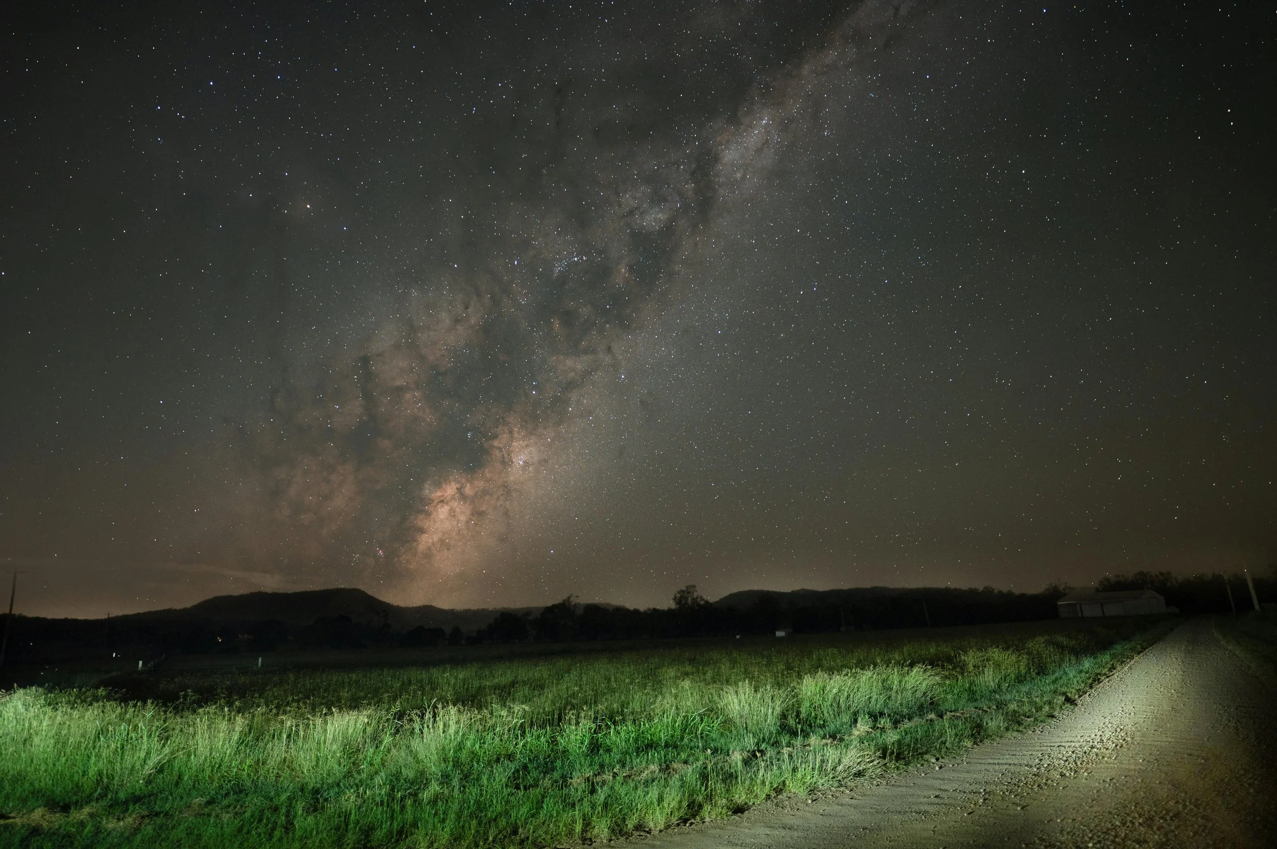 Clear night sky with Milky Way galaxy visible over a grassy field and dirt road.