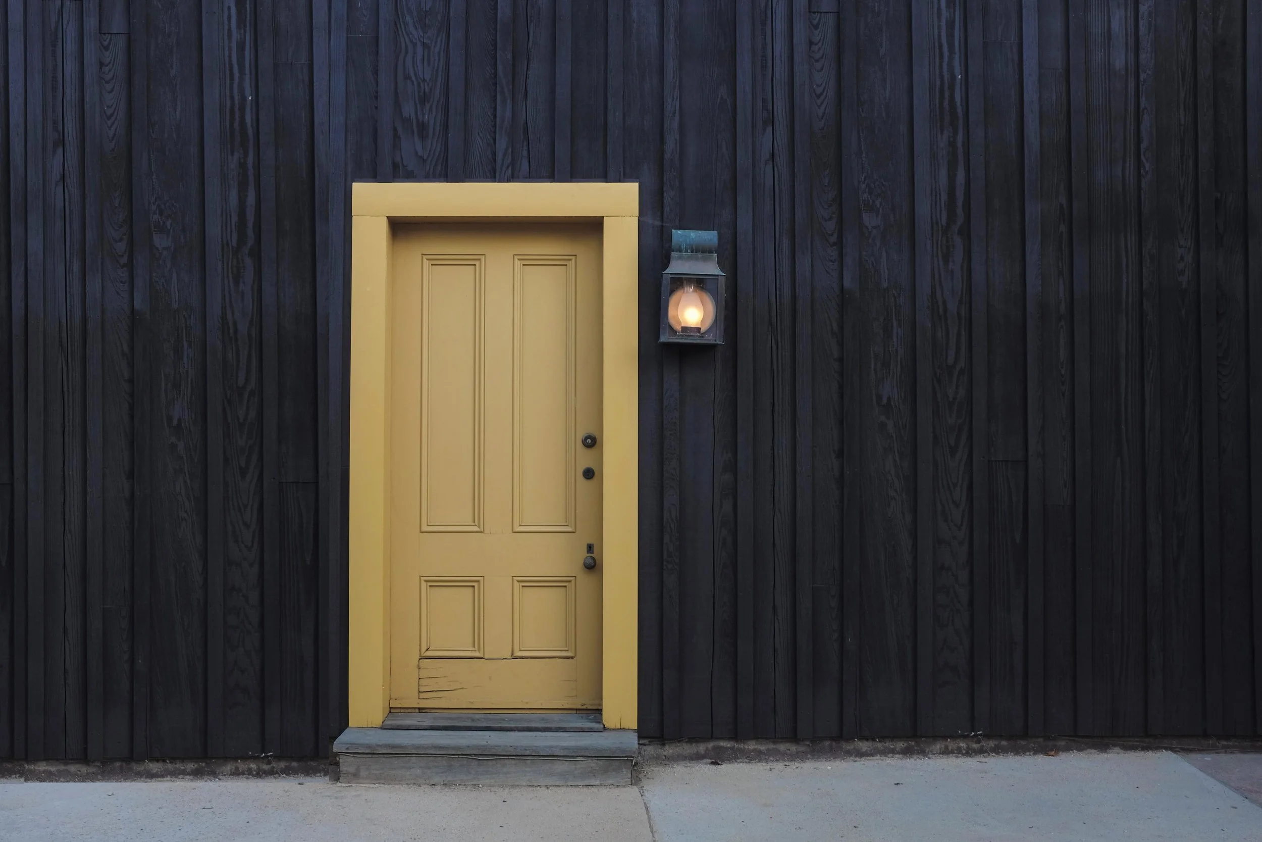 Yellow door on a black wooden wall with a lamp beside it.