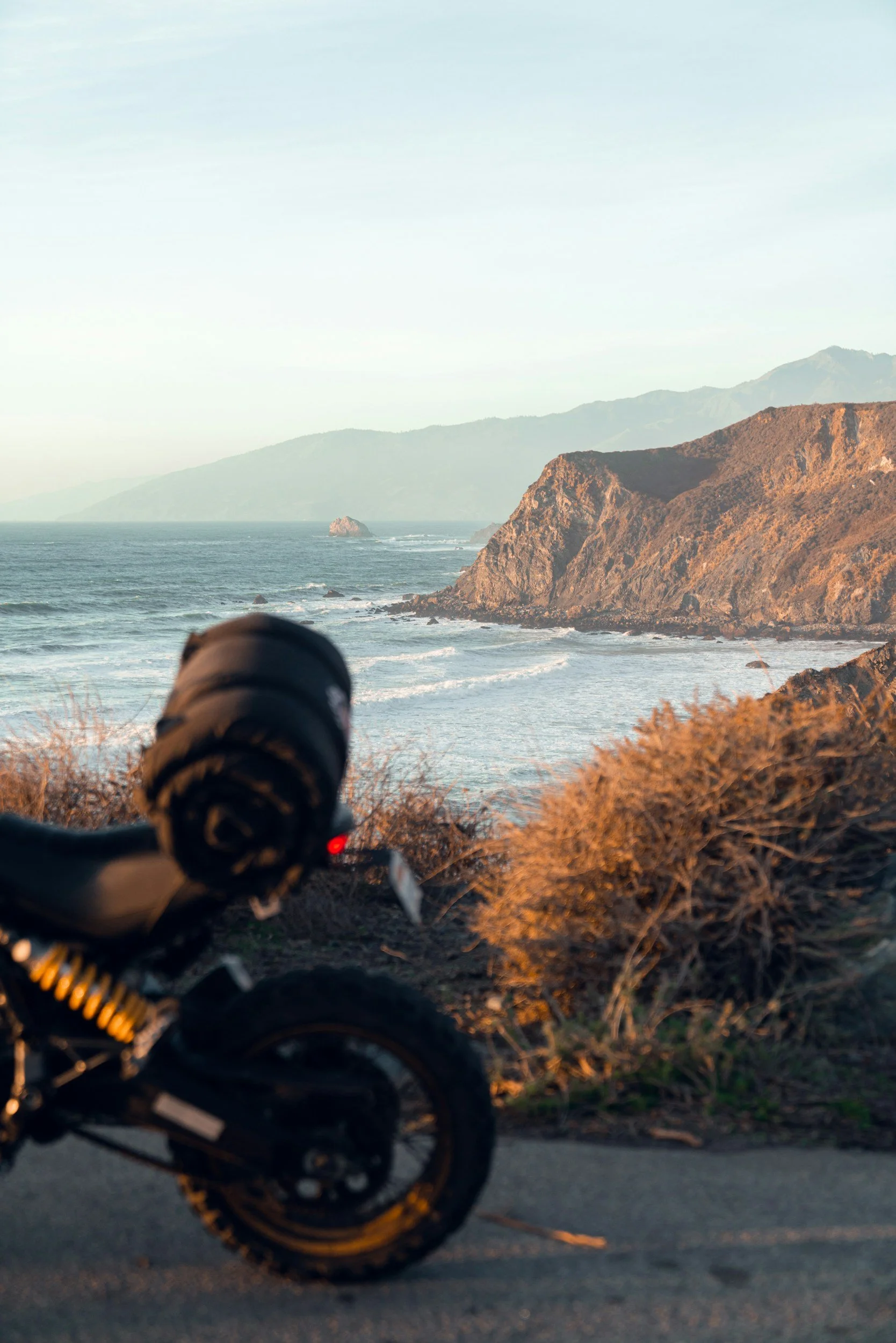touring motorcycle parked on a northern california coastal road with rocks, bushes, and ocean waves in the background, along with hills and trees.