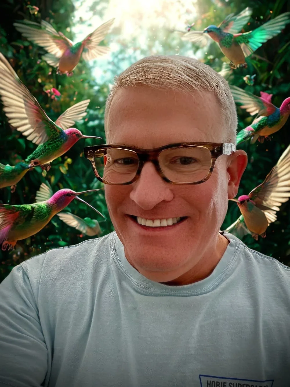 A smiling man with glasses taking a selfie in a lush, green setting surrounded by colorful hummingbirds flying around him.