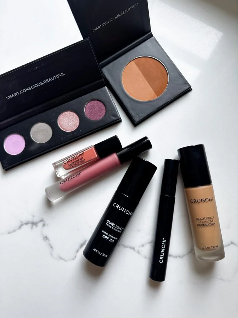 Makeup products including eyeshadow palettes, foundation, SPF moisturizer, liquid blush, and makeup brush on a marble surface.