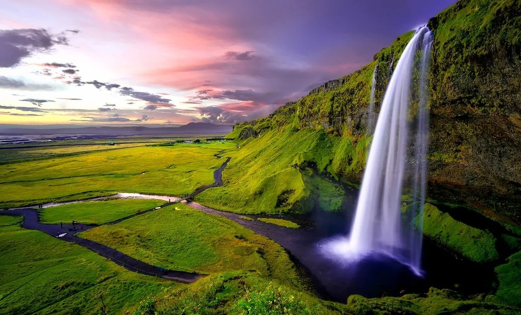 Long exposure of Seljalandsfoss waterfall cascading down a high, mossy green cliff under a vibrant pink and orange sunset sky.