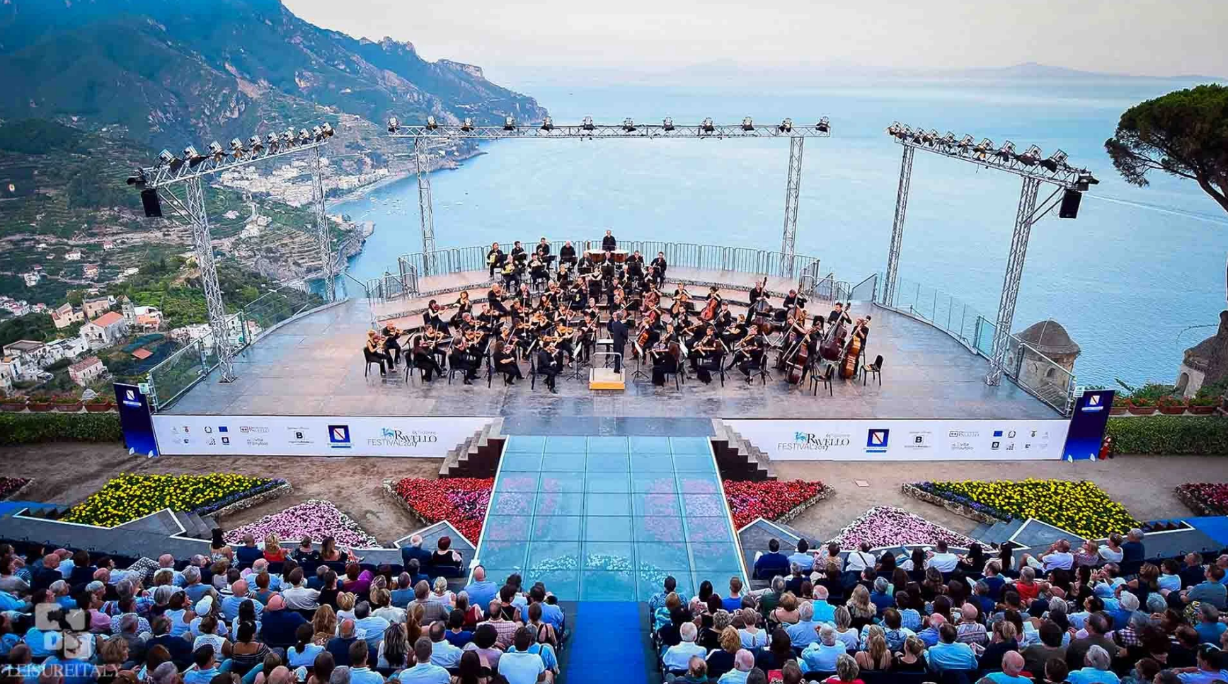 An orchestra performs on an outdoor stage in Ravello overlooking the sea and Amalfi Coast, with an audience in the foreground.