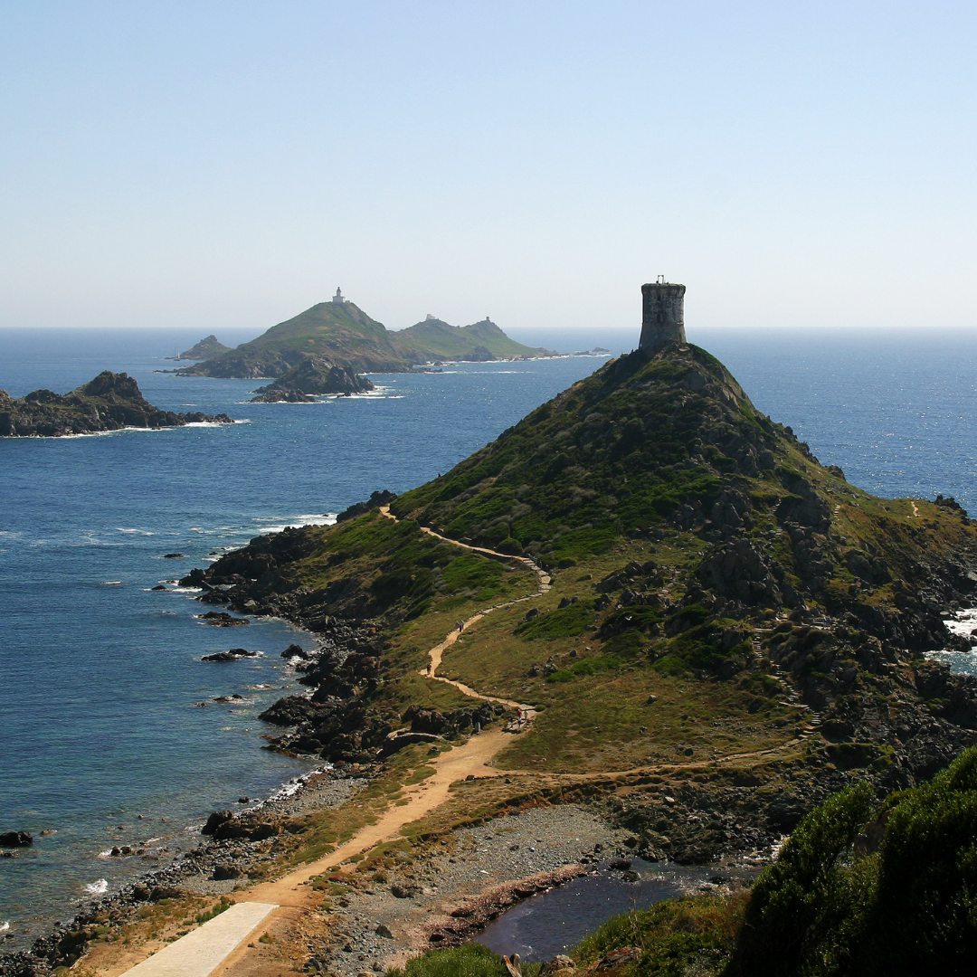 A stone Genoese watchtower perched atop a grassy green hill on a narrow peninsula surrounded by the blue sea.