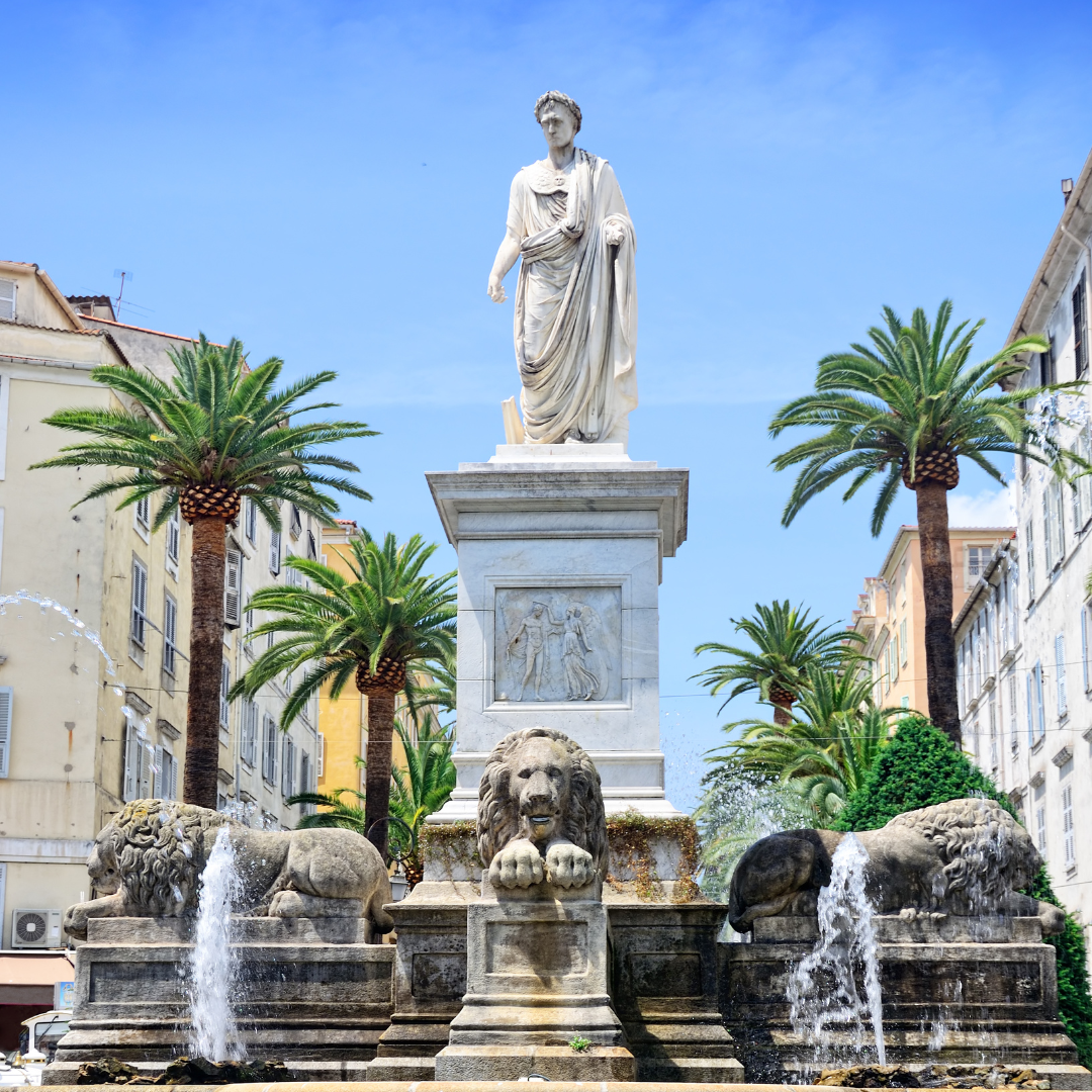 A white marble statue of Napoleon Bonaparte standing on a tall pedestal above carved lion fountains surrounded by palm trees.