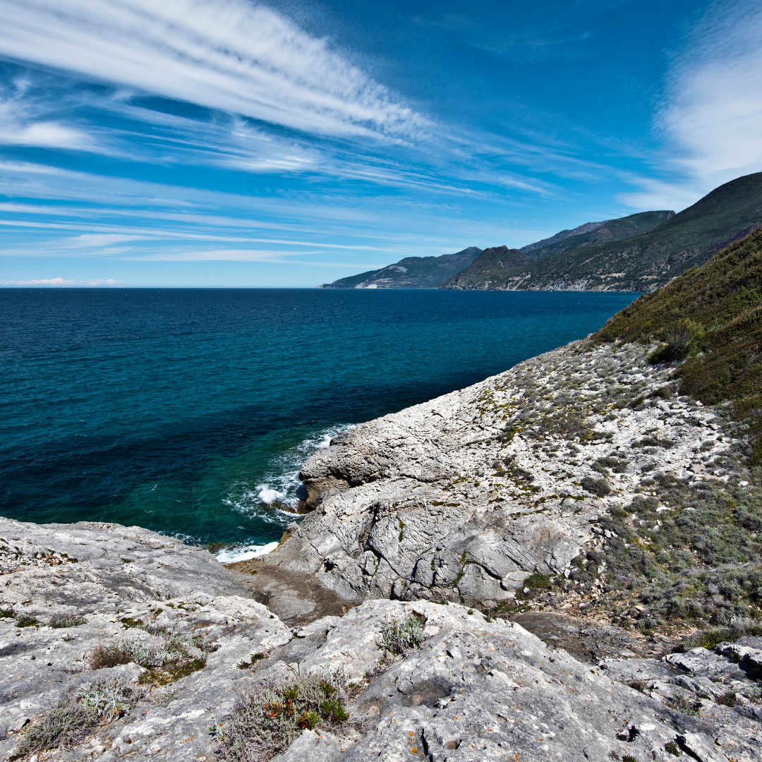 Rocky white coastal cliffs overlooking deep turquoise and royal blue Mediterranean ocean water in Corsica under a bright sky.