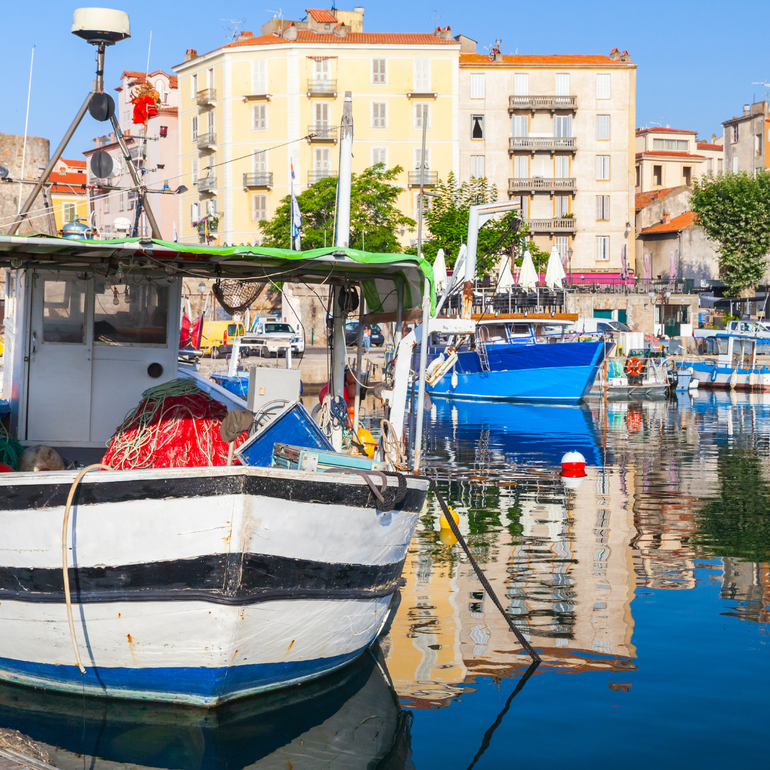 A wooden fishing boat docked in a calm harbor with reflections of yellow and pastel buildings on the water's surface.