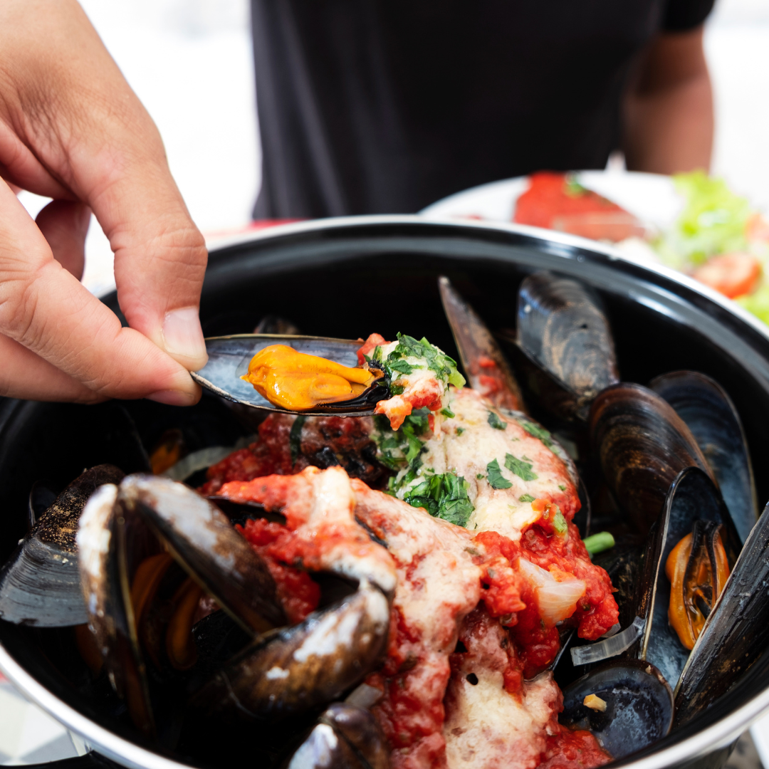 A hand picking a steamed mussel out of a black pot filled with fresh mussels cooked in tomato sauce and green herbs.