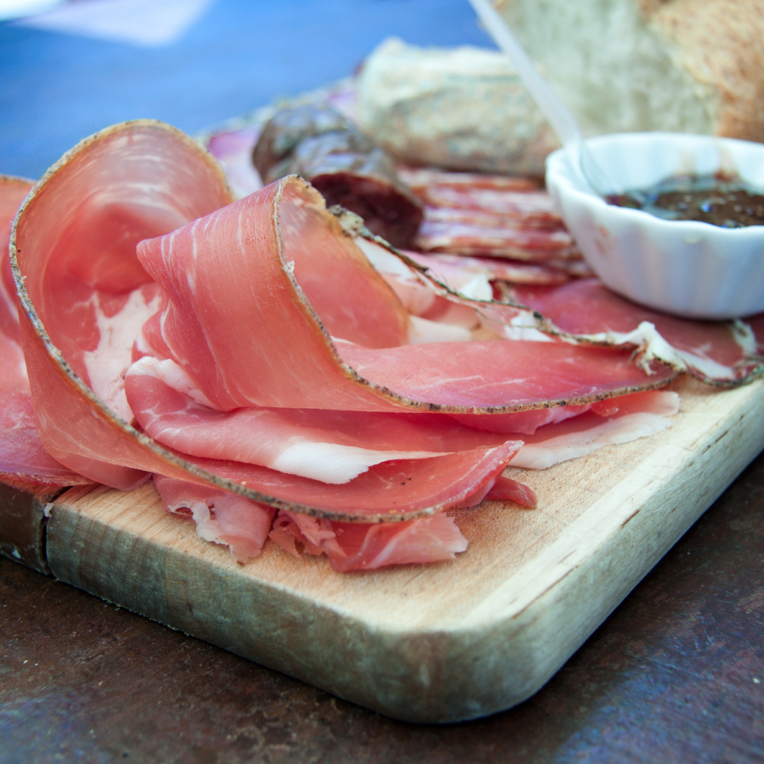 Sliced cured Corsican prosciutto ham arranged on a wooden serving board with rustic bread and a small bowl of fruit preserve.