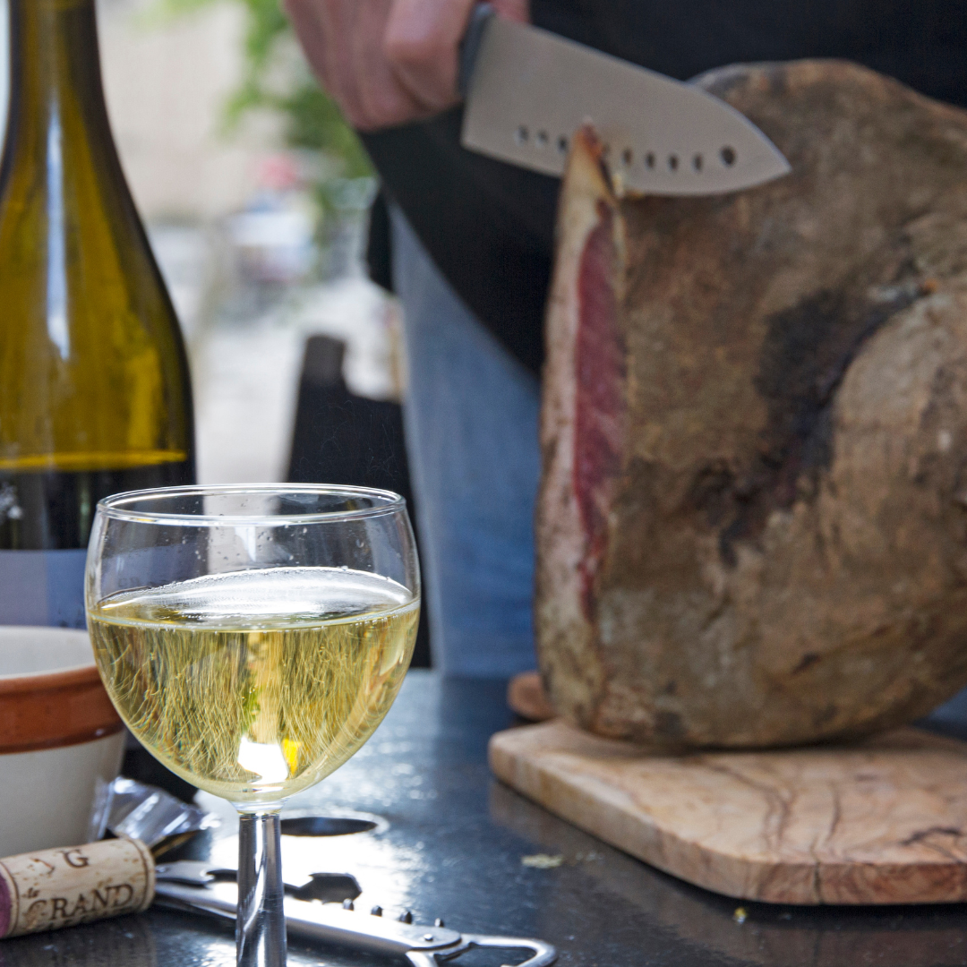A glass of crisp white wine on a table next to a chef hand-slicing a large cured leg of ham with a sharp knife.
