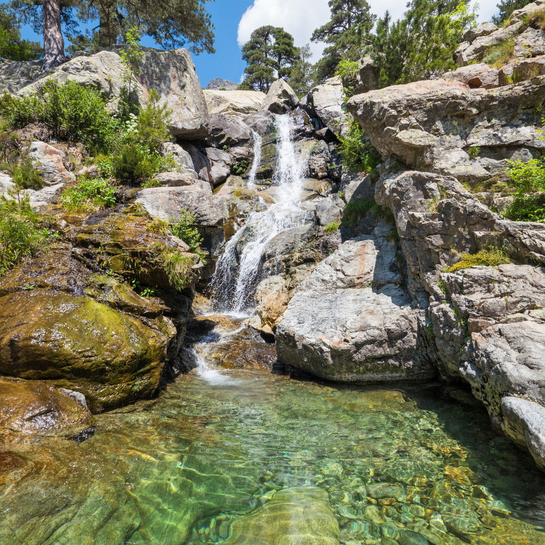 A freshwater mountain waterfall spilling over rocky granite ledges into a transparent, emerald-green swimming pool.