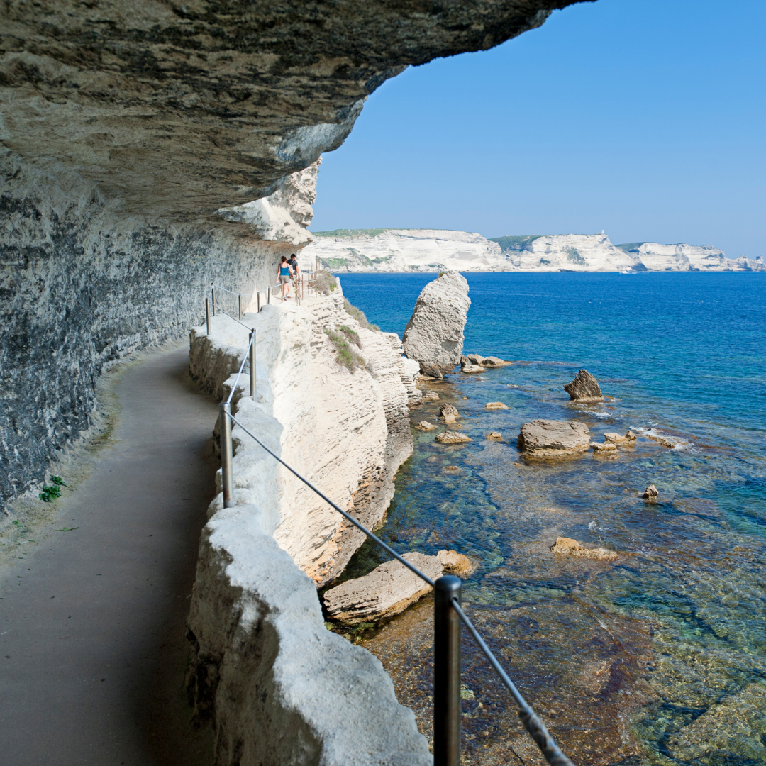 A narrow pedestrian walkway carved under a massive overhanging white cliff edge directly above clear blue sea water.