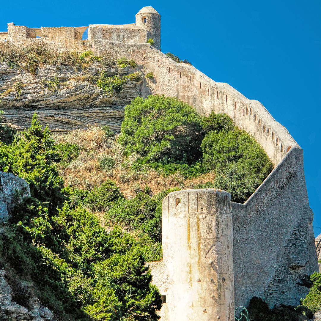 Low-angle view looking up at the thick stone fortification walls and watchtowers of an ancient Citadel built on a cliffside under a clear blue sky.