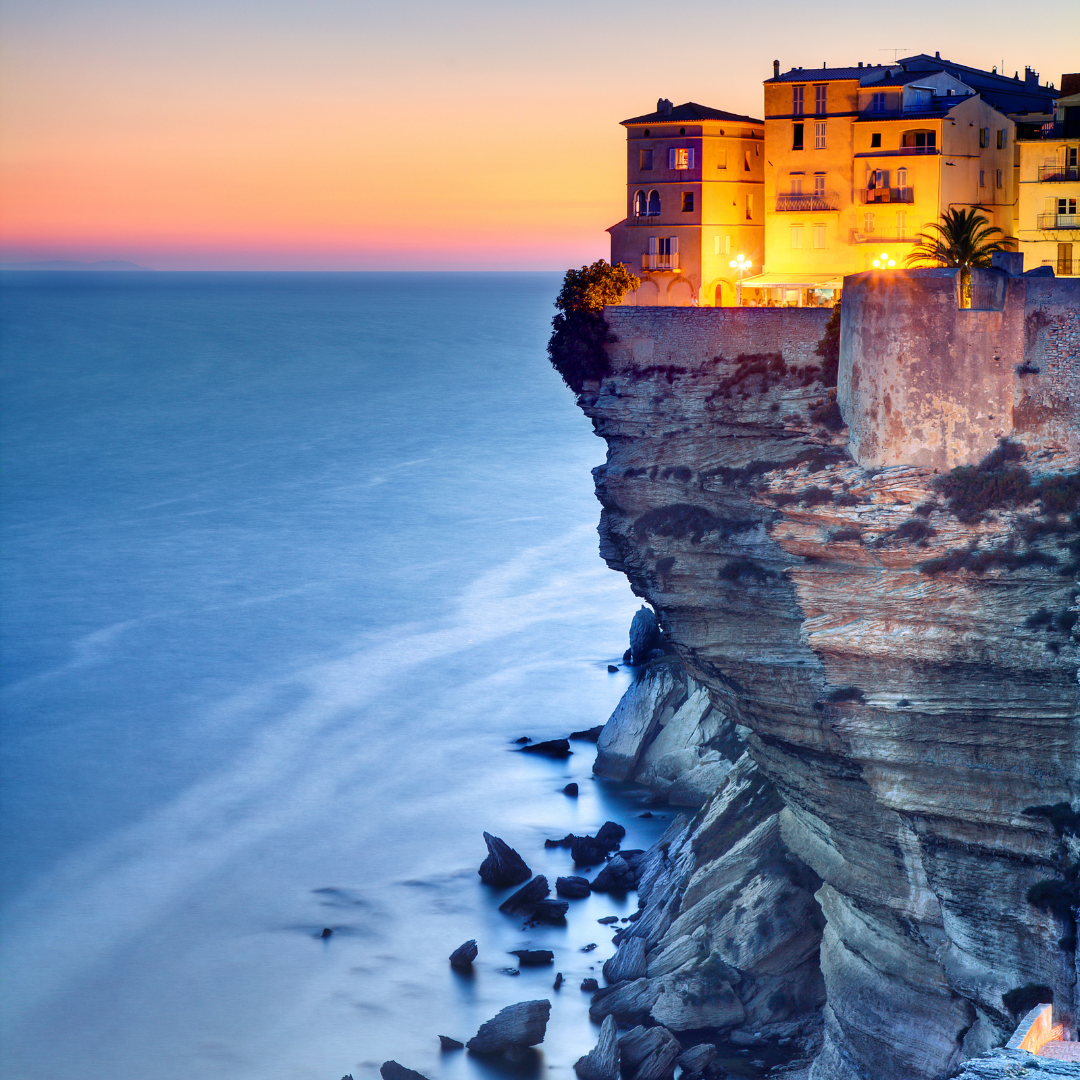Illuminated buildings resting on the edge of sheer white ocean cliffs at sunset overlooking calm blue waters.