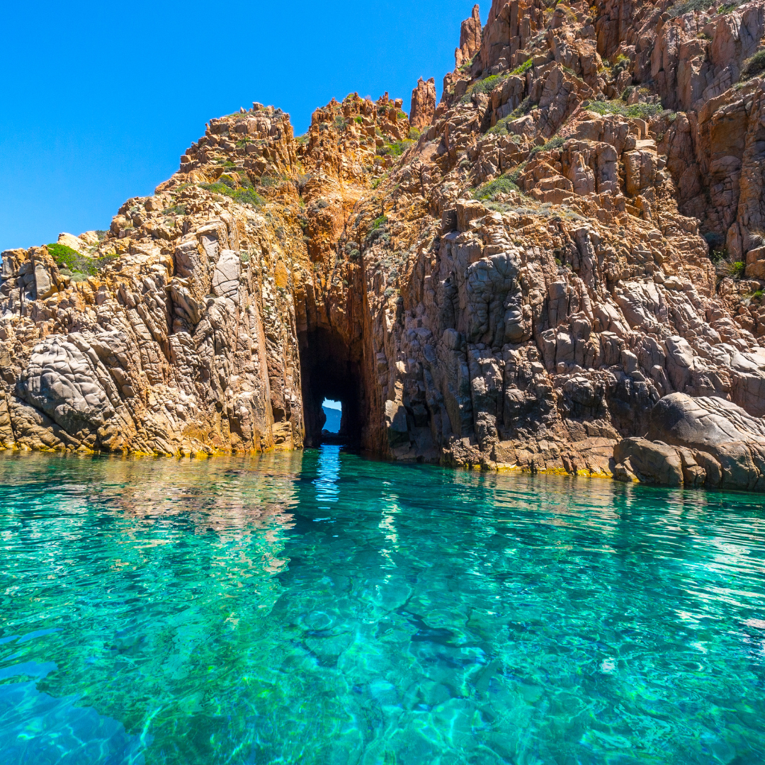 A natural stone archway sea cave opening through high, jagged red rock cliffs into clear, bright turquoise ocean water.